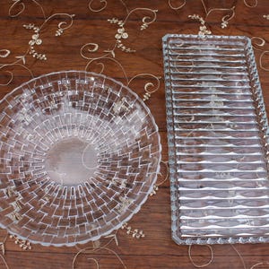 Puede incluir: Un cuenco de cristal transparente con una textura de tejido de cesta y una bandeja rectangular de cristal transparente a juego que presenta una serie de crestas horizontales elevadas. Ambos artículos son de cristal transparente y lisos.