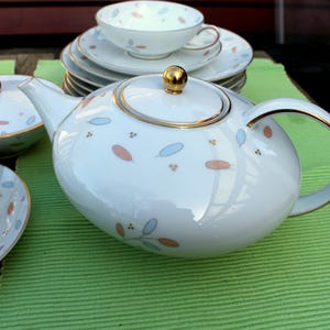 Könnte beinhalten: Weißes Porzellan-Teeservice mit Teekanne, Tassen, Untertassen und einer Schüssel mit Deckel. Das Set hat einen goldenen Rand und ein Muster aus blauen und pfirsichfarbenen blattartigen Designs. Die Teekanne hat einen goldenen Knopf auf dem Deckel.