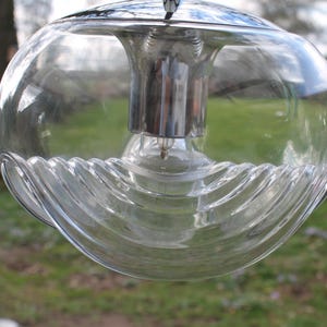 Può includere: Un lampadario in vetro trasparente con un paralume sferico e una parte superiore in metallo cromato. La parte inferiore del paralume presenta un design ondulato e strutturato. Il lampadario è appeso all'aperto con uno sfondo verde sfocato.