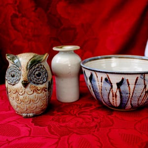 Può includere: Una statuetta di gufo in ceramica, un piccolo vaso bianco e una ciotola decorativa con disegni dipinti blu e marroni sono disposti su una superficie rossa a motivi. Il gufo ha dettagliate marcature di piume e grandi occhi. La ciotola ha un bordo largo.