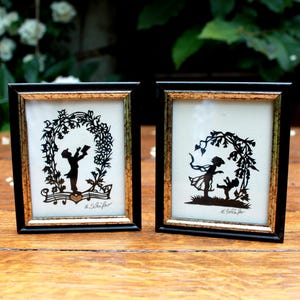 Peut inclure: Deux cadres photo rectangulaires aux bordures noires et dorées, contenant chacun une silhouette en papier noir. Le cadre de gauche montre un personnage tenant un livre sous une arche florale. Celui de droite représente deux personnages.