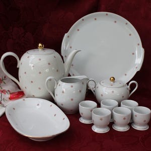 Op de afbeelding: Een wit porseleinen theeservies met een theepot, melkkan, suikerpot, serveerschaal, serveerschaal en eierdopjes. Elk stuk is versierd met kleine rode vierkante patronen en gouden accenten. Een bloemen servet is ook inbegrepen.