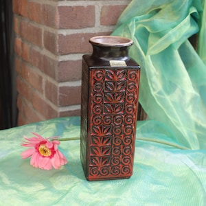 Può includere: Un vaso rettangolare in ceramica marrone scuro con un motivo geometrico rosso e nero. Il vaso ha un bordo leggermente svasato ed è decorato con un motivo ripetuto di foglie e volute stilizzate. Un fiore rosa con centro giallo è appoggiato vicino.