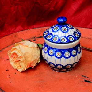 Può includere: Un zuccheriera in ceramica blu e bianca con coperchio, decorata con un motivo a piuma di pavone. La zucceriera è appoggiata su un vassoio rosso con una singola rosa color pesca.