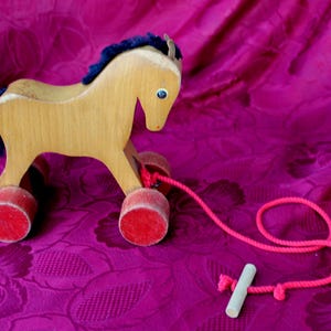 Può includere: Un cavallo di legno giocattolo con criniera e coda blu, su ruote rosse con uno spago rosa attaccato. Il cavallo è su uno sfondo di tessuto rosa.