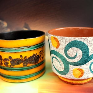 Könnte beinhalten: Zwei Keramik-Blumentöpfe mit bunten geometrischen Mustern. Ein Topf ist gelb mit grünen und schwarzen Streifen, der andere ist weiß mit grünen Wirbeln und gelben Akzenten.