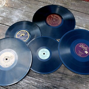 May include: A collection of vintage vinyl records, each with a dark blue surface and colorful labels. The records are arranged on a weathered wooden surface, showcasing their circular shape and aged appearance. The labels feature various fonts and designs.