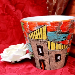 Könnte beinhalten: Eine handbemalte Keramiktasse mit einem Design aus stilisierten Gebäuden in Braun, Gelb, Blau und Rot. Die Tasse steht auf einer roten, gemusterten Oberfläche. Eine weiße Rose liegt in der Nähe.