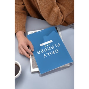 Peut inclure: Un agenda quotidien bleu avec le texte "DAILY PLANNER" en blanc. L'agenda a un petit rectangle blanc avec deux points jaunes sur le devant.