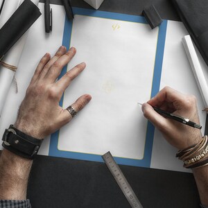 May include: A person is sketching on a white sheet of paper with a blue border. The paper has a ruler and the text "</>" at the top. The person is wearing a black leather bracelet on their left wrist and a brown leather bracelet with beads on their right wrist.