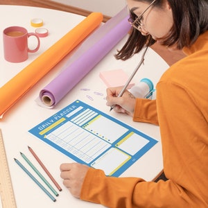 Peut inclure: Une personne &eacute;crit dans un agenda quotidien bleu avec un crayon. L'agenda a un design jaune et blanc. Il y a des crayons de couleur, une r&egrave;gle et une tasse rose sur la table.