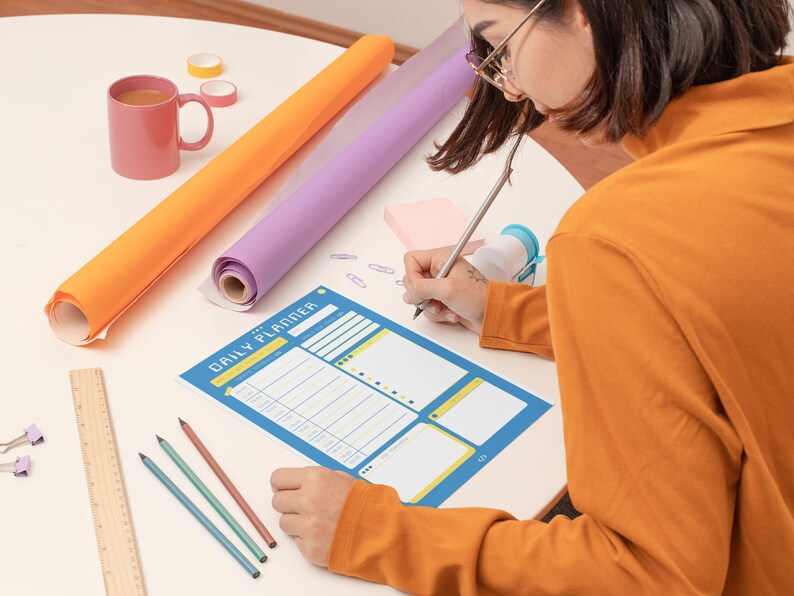 Peut inclure: Une personne &eacute;crit dans un agenda quotidien bleu avec un crayon. L'agenda a un design jaune et blanc. Il y a des crayons de couleur, une r&egrave;gle et une tasse rose sur la table.