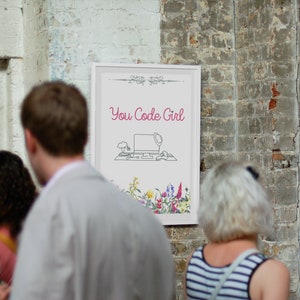 May include: A white framed poster with the text "You Code Girl" in pink lettering. The poster features a line drawing of a computer with a hat on top of it, a keyboard, and a mouse. The poster is surrounded by a border of flowers.