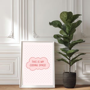 May include: A framed print with the text "THIS IS MY CODING SPACE" in a pink cloud shape. The background is light pink with small black dots. A large green plant in a dark basket sits to the right of the print.