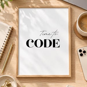 May include: A rectangular wooden frame containing a white print with the black text Time to CODE. The frame is positioned on a light wood surface alongside a laptop, a coffee cup, a smartphone, a gold pen, a spiral notebook, and binder clips.