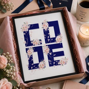 May include: A framed print featuring the word CODE in bold navy blue letters decorated with soft pink floral illustrations. The print is presented in a gift box alongside a navy card, a gold keychain, a notebook, and a mug with text.