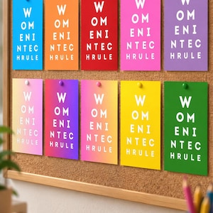 May include: A cork bulletin board displays ten rectangular cards in various solid and gradient colors. Each card features the white text "WOMEN IN TECH RULE" arranged in five stacked lines. The cards are secured to the board with small push pins.