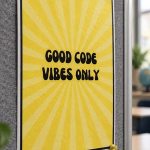 Op de afbeelding: Een rechthoekige poster met een geel zonnestraalpatroon en een zwarte rand. De tekst "GOOD CODE VIBES ONLY" staat in het midden gedrukt in een vet, zwart, afgerond lettertype. De poster is met vier gekleurde punaises aan een grijze wand bevestigd.