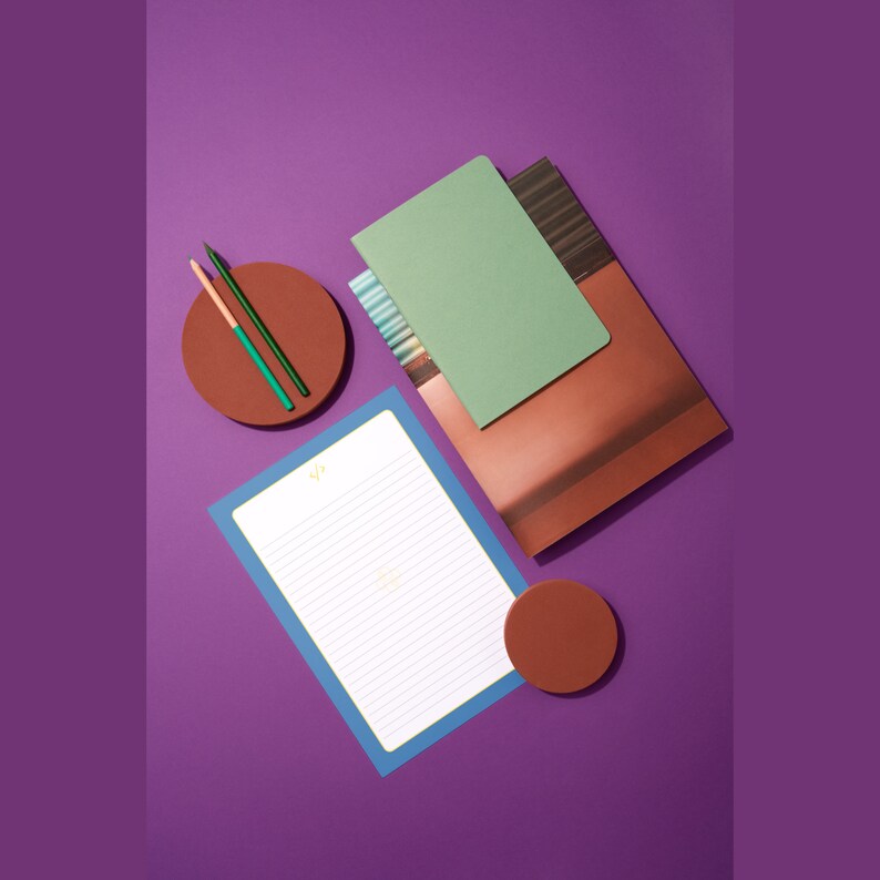 May include: A flat lay of stationery supplies on a purple background. There are two green pencils, a brown and green notebook, a brown and green journal, a white lined notepad, and two brown coasters.