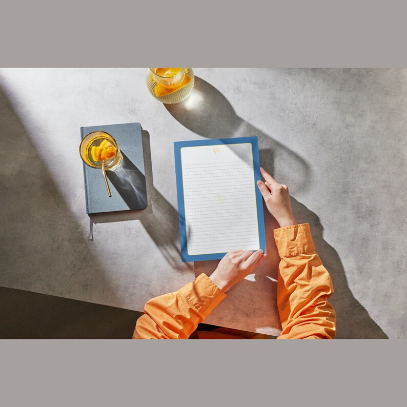 May include: A person holding a blue lined notepad with a gold logo. The notepad is on a gray surface with a glass of orange juice and a gray notebook.