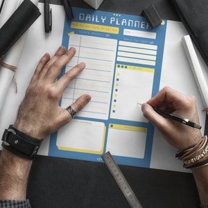 Peut inclure: Un planificateur quotidien bleu et jaune avec des sections pour l'horaire du jour, les objectifs du jour, la liste des choses &agrave; faire, pour demain, les notes et une r&egrave;gle. Une personne &eacute;crit sur le planificateur avec un stylo noir.