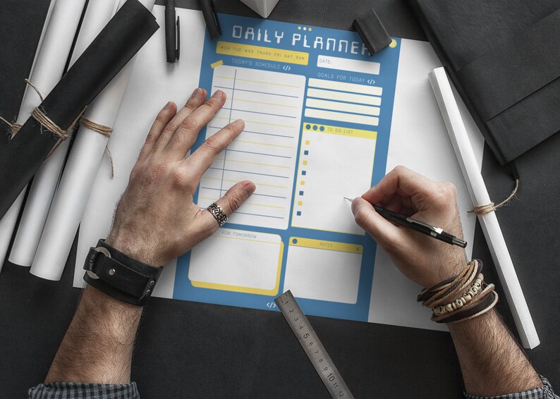 Peut inclure: Un planificateur quotidien bleu et jaune avec des sections pour l'horaire du jour, les objectifs du jour, la liste des choses &agrave; faire, pour demain, les notes et une r&egrave;gle. Une personne &eacute;crit sur le planificateur avec un stylo noir.