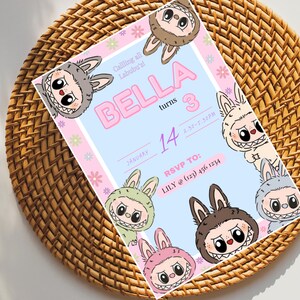 May include: A pink and blue birthday invitation with a cartoon character on the front. The invitation says "Calling all Labubu's! Bella turns 3 January 14 3:30-5:30PM RSVP TO: LILY @ (123) 456 1234"