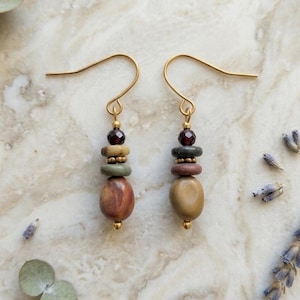 May include: A pair of dangle earrings featuring gold-toned ear wires, small dark red faceted beads, and a stack of smooth, multi-colored natural stone beads in shades of tan, sage green, and reddish-brown, suspended from a simple metal hook.