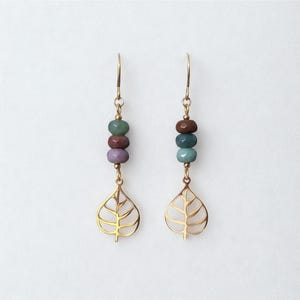 Può includere: Un paio di orecchini color oro con un ciondolo a forma di foglia. Ogni orecchino presenta tre perline colorate nelle tonalità del verde, marrone e viola. Gli orecchini sono sospesi a un gancio color oro.