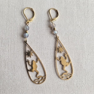 Può includere: Un paio di orecchini color oro. Ogni orecchino presenta un pendente a forma di goccia con un design traforato di una rana, stelle e acqua. Gli orecchini hanno una chiusura a leva e sono impreziositi da tre piccole perle azzurre.