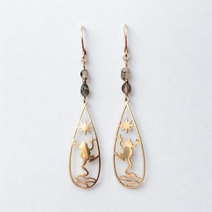 Può includere: Un paio di orecchini pendenti color oro. Ogni orecchino presenta un design a goccia con un ritaglio di una rana, un sole e acqua. Gli orecchini sono adornati con piccole perline grigie e marroni.