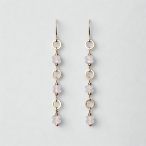 Può includere: Un paio di orecchini pendenti color oro. Ogni orecchino presenta un design delicato con piccole pietre preziose rosa chiaro e anelli color oro. Gli orecchini hanno una semplice chiusura a gancio.