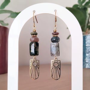 May include: Dangling earrings with a gold-coloured sunburst charm. The earrings feature natural stone beads in green, brown, and grey. The earrings are suspended from gold-coloured ear wires.