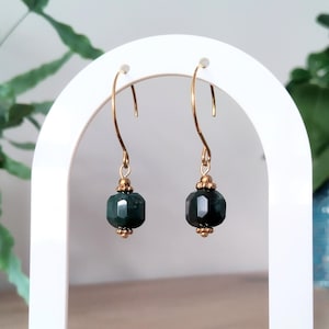 Peut inclure: Une paire de boucles d'oreilles pendantes avec des perles vert foncé facettées suspendues à des crochets dorés. Les perles sont accentuées de petites perles dorées. Les boucles d'oreilles sont présentées sur un présentoir blanc.