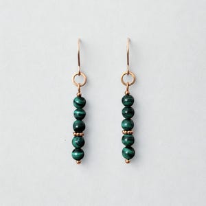 Peut inclure: Une paire de boucles d'oreilles pendantes avec des perles de malachite vert foncé. Les perles sont enfilées sur un fil de couleur or, avec un petit anneau de couleur or en haut et une perle de couleur or en bas. Sur fond blanc.