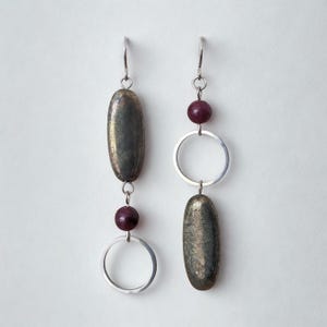 May include: A pair of dangle earrings. Each earring has a silver hook, a dark metallic oval bead, a small round purple bead, and a silver hoop. The earrings are set against a white background.