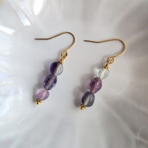 Könnte beinhalten: Ein Paar Ohrhänger mit goldfarbenen Haken und jeweils drei facettierten Perlen. Die Perlen zeigen einen Farbverlauf aus durchscheinenden violetten, lavendelfarbenen und blassgrünen Tönen. Jeder Ohrring endet mit einer kleinen goldfarbenen Perle.