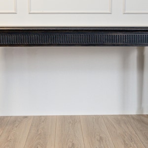 Puede incluir: Una consola negra con una tapa rectangular y cuatro patas torneadas. La mesa presenta un diseño tallado detallado a lo largo del borde superior. La mesa es de madera y tiene un acabado mate. La mesa mide aproximadamente 152 cm de largo.