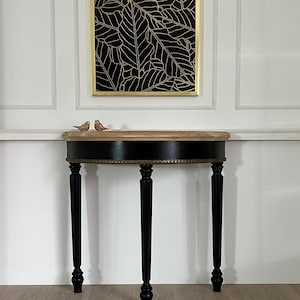 Può includere: Un tavolino nero e oro con piano rotondo e quattro gambe nere. Il tavolo ha un bordo dorato attorno al piano. Sopra il tavolo c'è un disegno di foglie nere e oro incorniciato. Due piccole statuette di uccelli sono sul tavolo.