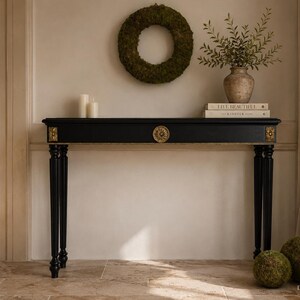Op de afbeelding: Een zwarte sidetable met vier slanke poten en goudkleurige decoratieve accenten op de hoeken en in het midden. De tafel heeft een rechthoekig blad en is versierd met een cirkelvormig gouden medaillon met gedetailleerde afwerking.