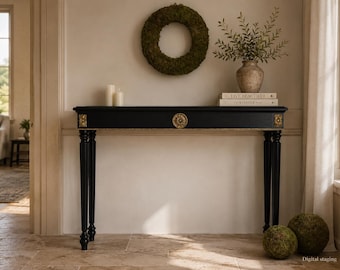 May include: A black console table with four slender legs and gold-toned decorative accents on the corners and centre. The table features a rectangular top and is adorned with a circular gold medallion. The design includes intricate detailing.
