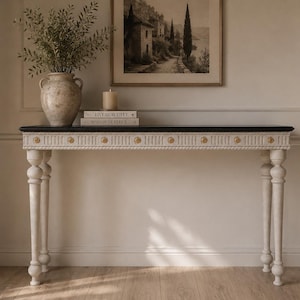 May include: A rectangular console table with a black top and a white base featuring fluted detailing, rope-style trim, and small gold-toned circular accents along the front edge. The table stands on four turned legs with a distressed white finish.