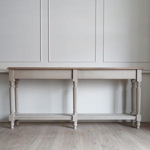 Può includere: Un tavolo consolle di colore grigio chiaro con un piano in legno e un ripiano inferiore. Il tavolo ha quattro gambe tornite con dettagli scanalati.