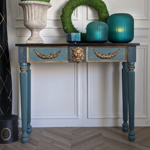 May include: A teal blue console table with gold accents and lion head carvings. The table has two drawers and sits on four fluted legs. The table is decorated with a wreath, two glass lanterns, and a small vase.