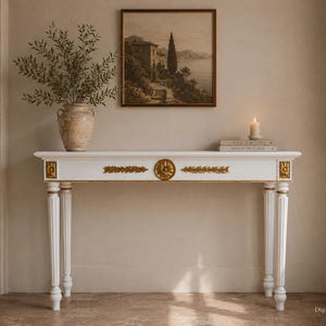 May include: A white console table featuring gold-toned decorative carvings along the front apron and fluted legs. The rectangular table has a flat top surface and is accented with ornate circular and linear gold leaf-style embellishments.