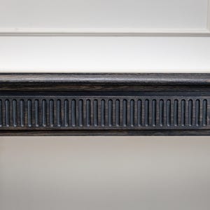 Puede incluir: Primer plano de una moldura de madera gris oscuro con un patrón ranurado vertical detallado. La moldura forma parte de un elemento arquitectónico más grande, sobre una pared blanca con una moldura simple.