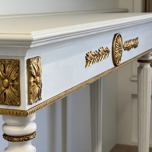 Op de afbeelding: Een witte console met gouden accenten. De tafel heeft gouden bladversieringen aan de voorkant, waaronder rechthoekige en cirkelvormige decoratieve elementen. De poten zijn wit met gouden randen.