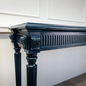 Puede incluir: Una consola azul marino con detalles tallados ornamentados. La mesa presenta una parte superior rectangular, detalles tallados decorativos y detalles de listones verticales. La mesa está sostenida por dos patas estriadas. Las dimensiones de la mesa no son visibles.