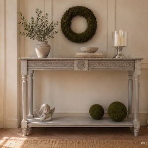 May include: A rectangular console table with a distressed grey finish, featuring fluted legs and intricate carved detailing along the apron. The table includes a lower shelf holding a small angel figurine and two moss spheres, with a wreath above.