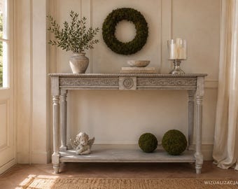 May include: A rectangular console table with a distressed grey finish, featuring fluted legs and intricate carved detailing along the apron. The table includes a lower shelf holding a small angel figurine and two moss spheres, with a wreath above.
